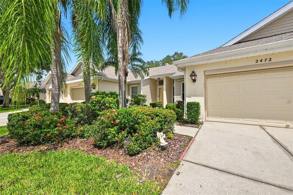 A beige house exterior features a two-car garage, a concrete driveway, and lush landscaping — 2472 NANTUCKET HARBOR LOOP, SUN CITY CENTER, FL