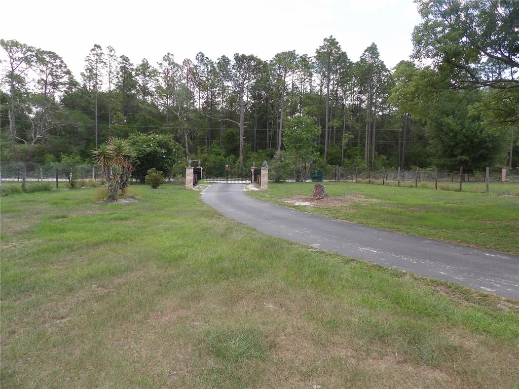 A paved driveway leads to a gated entrance on a grassy lot with surrounding trees — 18211 OAKLAWN, SPRING HILL, FL
