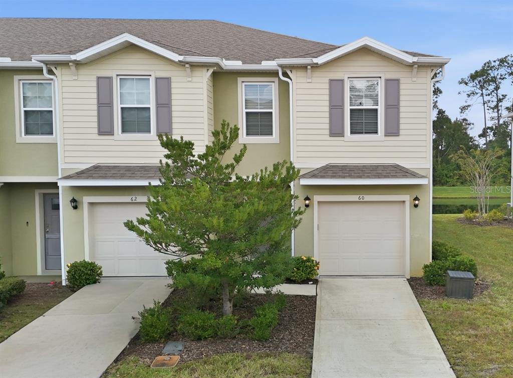 A two-story townhouse exterior features beige siding, two garage doors, and a central tree — 60 BELLA LAGO CIRCLE, PORT ORANGE, FL