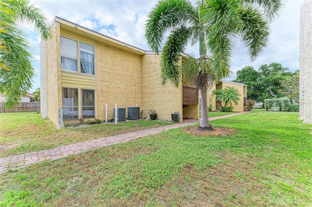 The exterior of a two-story yellow stucco apartment building with a brick walkway and palm trees — 3471 CLARK ROAD UNIT 169, SARASOTA, FL