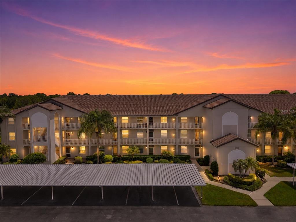 An exterior view of a multi-story apartment building at sunset with a covered parking area — 8735 OLDE HICKORY AVENUE UNIT 8304, SARASOTA, FL
