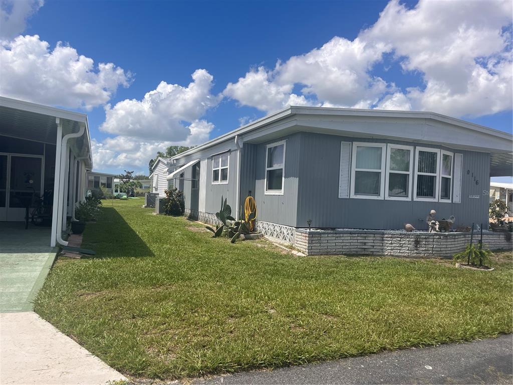 A gray manufactured home with a brick skirt sits on a grassy lot under clouds — 5316 53RD AVENUE E UNIT Q116, BRADENTON, FL