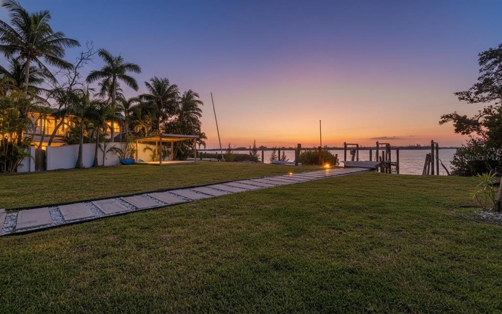 A grassy backyard features a stone path leading to a wooden dock at sunset — 10218 46TH AVENUE W, BRADENTON, FL