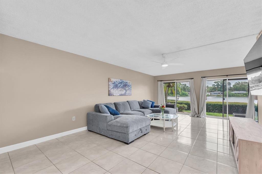 A living room features a gray sectional sofa, tile flooring, and sliding glass doors overlooking water — 1655 S HIGHLAND AVENUE UNIT J194, CLEARWATER, FL