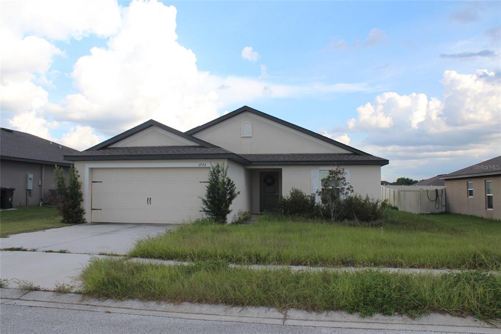 A single-story house with a beige stucco exterior, a two-car garage, and an overgrown lawn — 1733 CANAAN LOOP, DUNDEE, FL