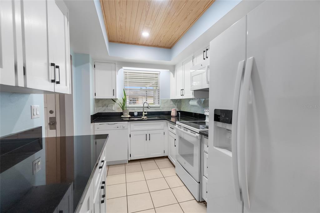 A galley kitchen features white cabinets, black countertops, a wood-paneled ceiling, and tile flooring — 676 PARK CIRCLE, BRADENTON, FL