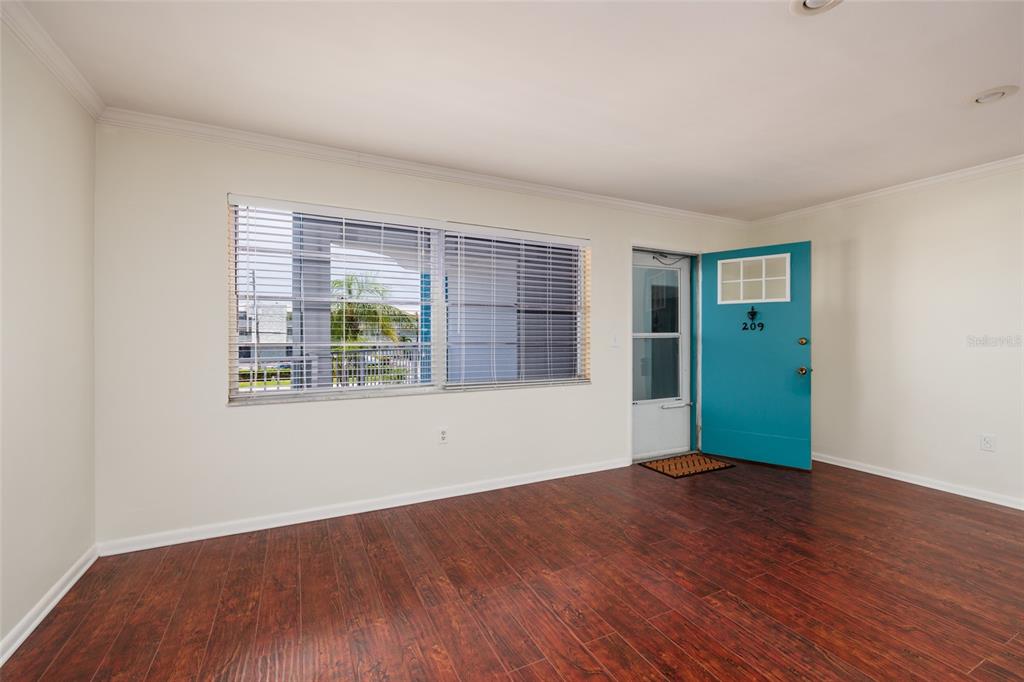 An empty room features wood-look flooring, a large window with blinds, and a teal door — 1893 SHORE DRIVE S UNIT 209, SOUTH PASADENA, FL
