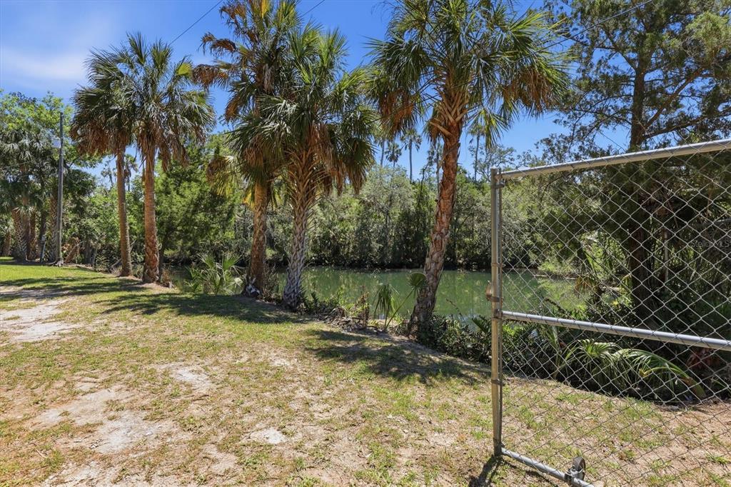 A chain-link fence stands beside a grassy yard with palm trees overlooking a small canal — 6240 COLONY CIRCLE, WEEKI WACHEE, FL