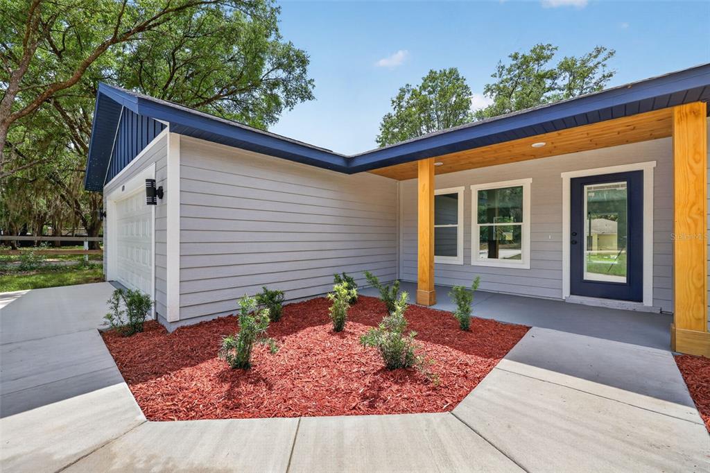 A gray house exterior features a blue door, wood support posts, and a red mulch garden — 13932 SW 104TH LANE, DUNNELLON, FL