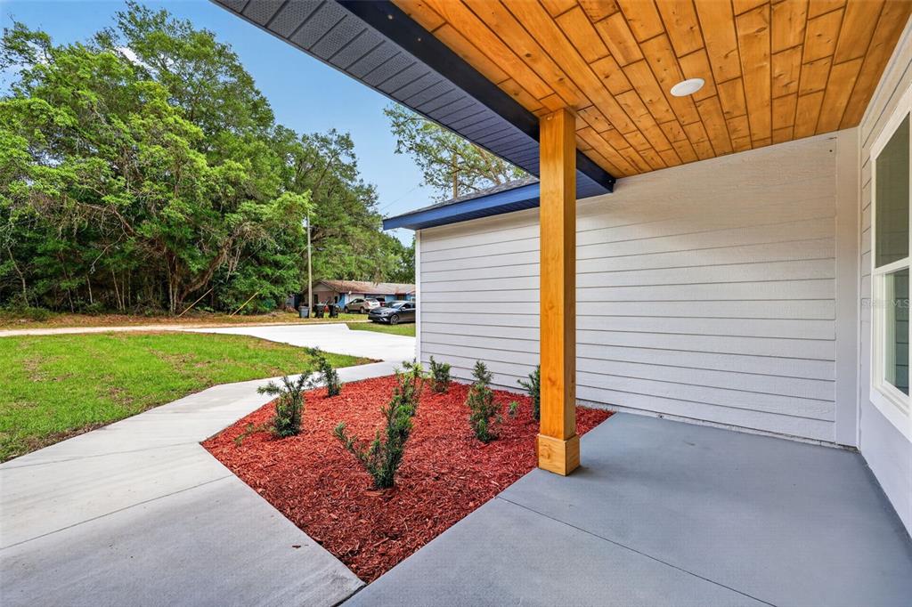 A covered concrete porch with a wood-paneled ceiling, a wooden post, and a landscaped bed — 13932 SW 104TH LANE, DUNNELLON, FL