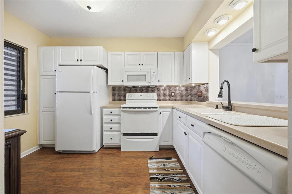 A kitchen features white cabinets, white appliances, a tiled backsplash, and wood-look flooring — 1802 MARINER DRIVE E UNIT 25, TARPON SPRINGS, FL