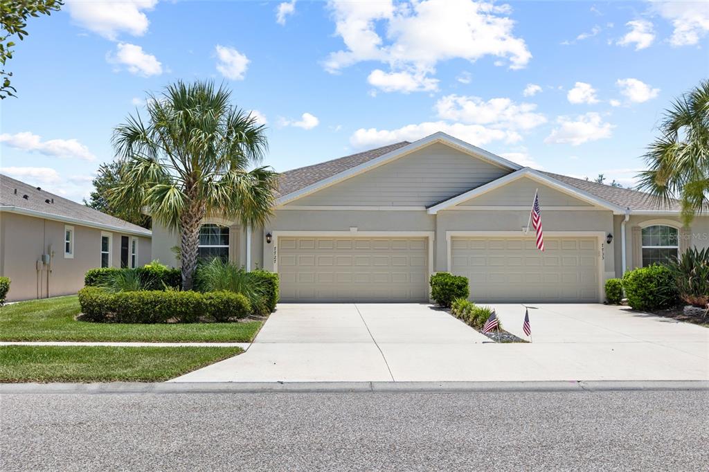 A duplex home with two garage doors, beige siding, and a palm tree in the yard — 7727 TIMBERVIEW LOOP, WESLEY CHAPEL, FL