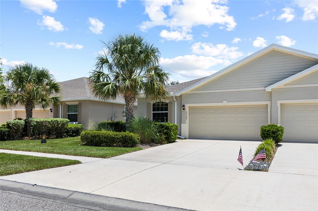 A beige house with a two-car garage, palm tree, and manicured landscaping under a blue sky — 7727 TIMBERVIEW LOOP, WESLEY CHAPEL, FL