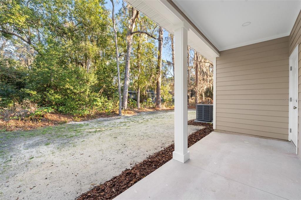 A covered concrete patio overlooks a sandy backyard area with trees and an air conditioner — 2016 NE 3RD AVENUE, GAINESVILLE, FL