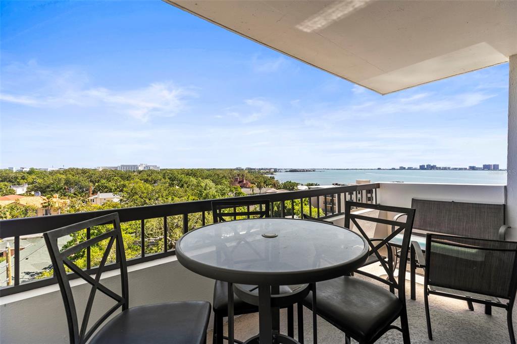A balcony with a glass-topped table and chairs overlooks a bay and surrounding trees — 30 TURNER STREET UNIT 704, CLEARWATER, FL