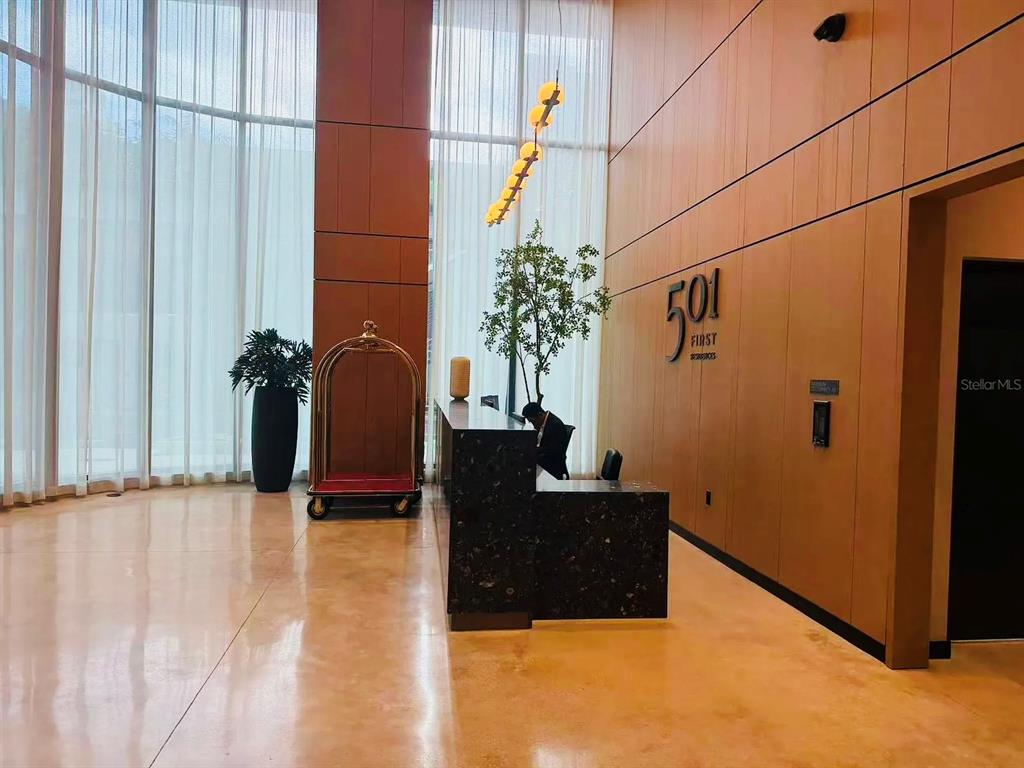 A modern building lobby features a dark stone reception desk, wood-paneled walls, and floor-to-ceiling windows — 121 NE 5TH STREET UNIT 1500, MIAMI, FL