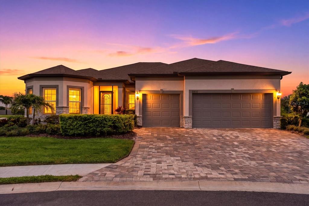 A single-story house with a paved driveway, three-car garage, and a sunset sky background — 8510 COASTAL PALMS GLEN, PARRISH, FL