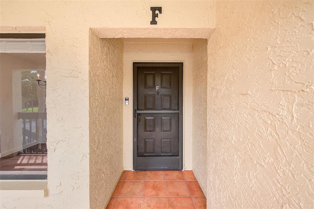 A dark brown storm door is centered in a stucco entryway with orange tile flooring — 1801 E LAKE ROAD UNIT 10F, PALM HARBOR, FL