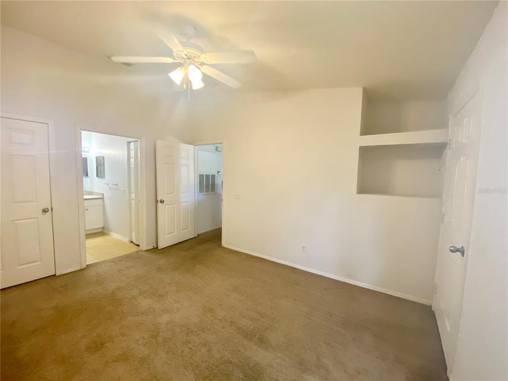 A bedroom with beige carpet, a ceiling fan, a bathroom doorway, and built-in wall shelving — 36101 DEER CREEK DRIVE UNIT 202, ZEPHYRHILLS, FL