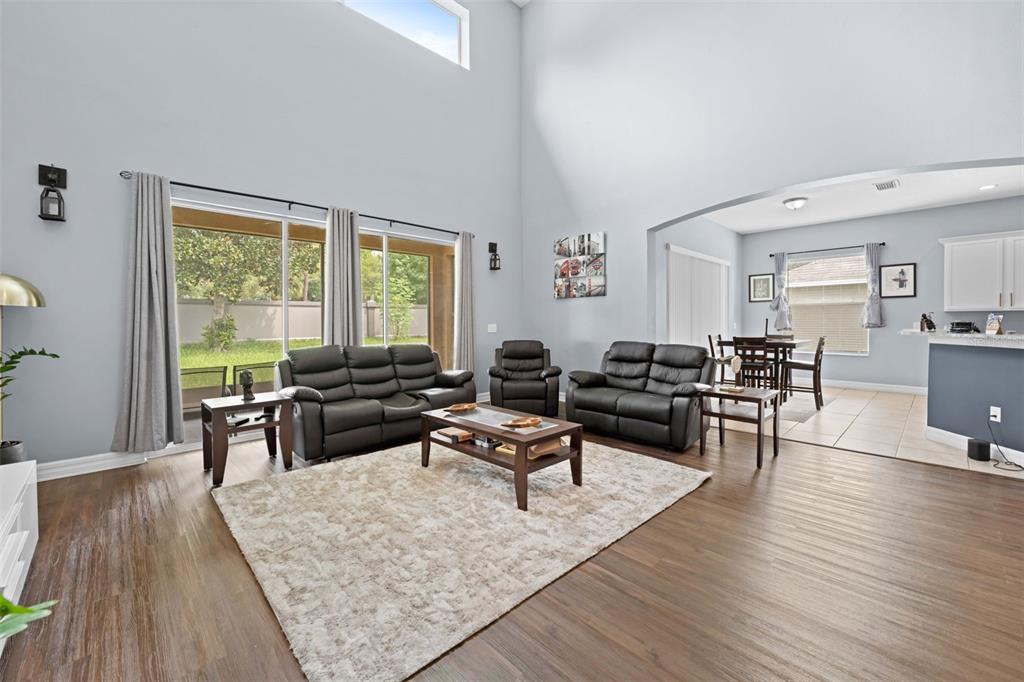 A living room with high ceilings, wood-look flooring, and black leather furniture sits near a dining area — 4501 BIRCHFIELD LOOP, SPRING HILL, FL