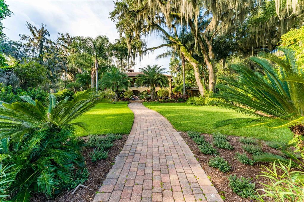 A brick walkway leads through a lush, tree-filled yard toward a two-story house — 8289 COLEE COVE ROAD, ST AUGUSTINE, FL