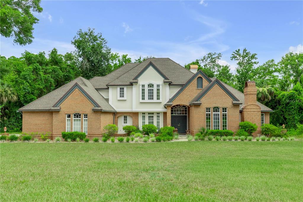 A two-story brick house with a large lawn and surrounding trees under a blue sky — 18 LOST CREEK LANE, ORMOND BEACH, FL