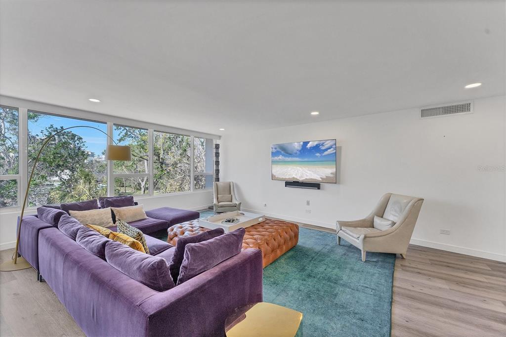 A living room features a purple sectional, a leather ottoman, and large windows overlooking trees — 100 SANDS POINT ROAD UNIT 323, LONGBOAT KEY, FL