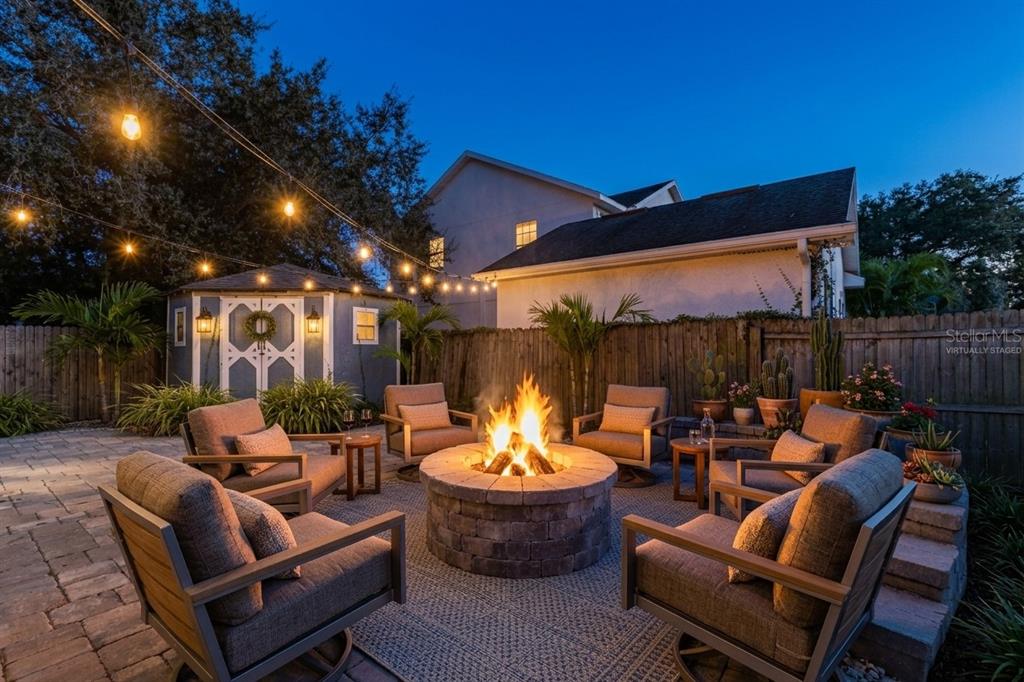 A backyard patio features a stone fire pit surrounded by chairs under string lights at dusk — 3208 W NAPOLEON AVENUE, TAMPA, FL