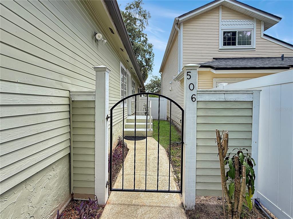 A black metal gate stands between two light green siding walls leading to a walkway — 506 HARVARD PLACE, APOPKA, FL