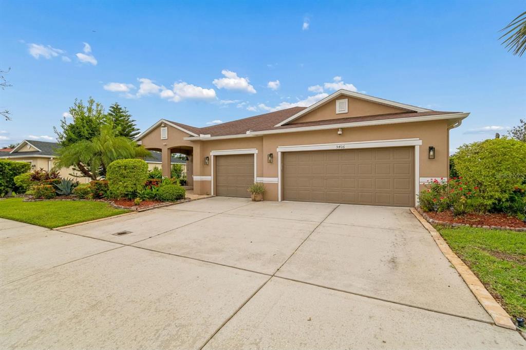 A tan single-story house features a three-car garage and a concrete driveway under a blue sky — 5406 SANDY SHELL DRIVE, APOLLO BEACH, FL
