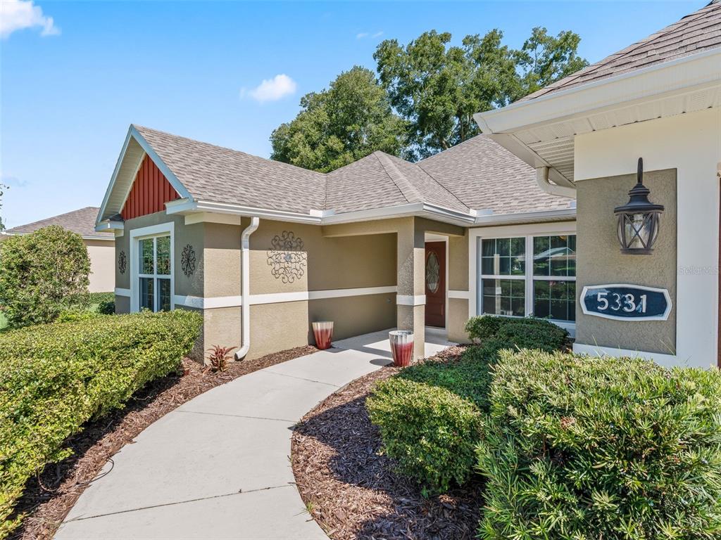 A concrete walkway leads to the front entrance of a tan house with a porch — 5331 GROVE MANOR, LADY LAKE, FL