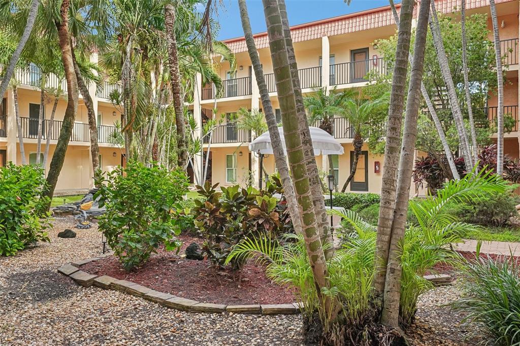 A courtyard with palm trees, tropical landscaping, and a multi-story apartment building with balconies — 8911 BLIND PASS ROAD UNIT 104, ST PETERSBURG, FL