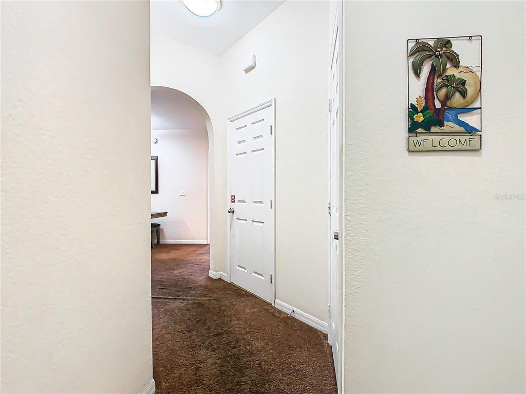A hallway with brown carpet leads to an arched doorway, featuring a white door and wall decor — 3050 PIRATES RETREAT COURT UNIT 108, KISSIMMEE, FL