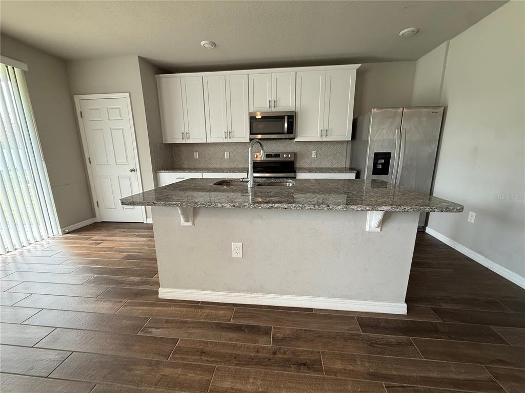 A kitchen features a granite-topped island, white cabinetry, stainless steel appliances, and wood-look tile flooring — 5264 STEPHS FIELD STREET, ST CLOUD, FL