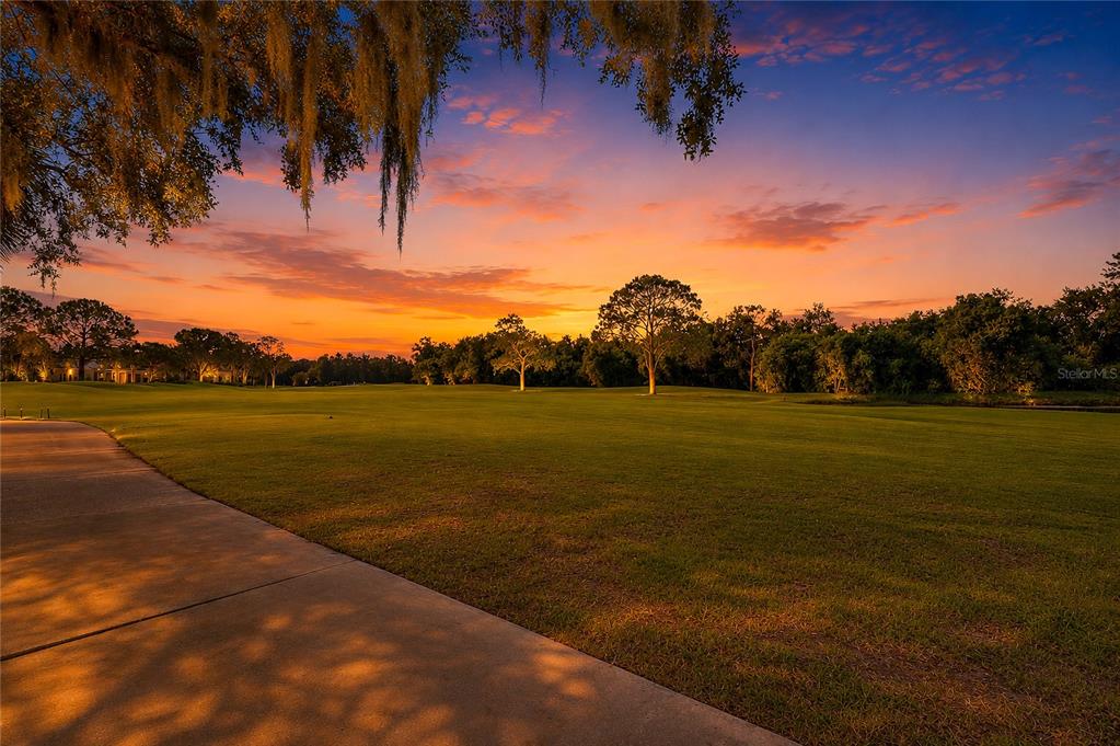 A paved path leads toward a grassy golf course under a vibrant sunset sky — 8718 54TH AVENUE E, BRADENTON, FL