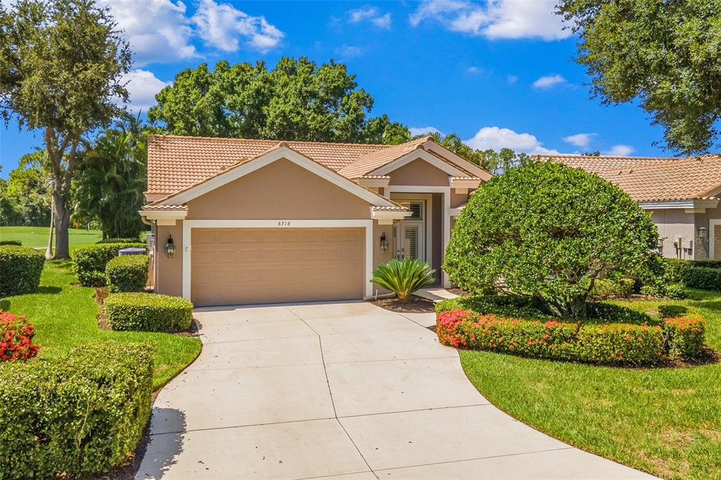A tan single-story house features a two-car garage, a concrete driveway, and landscaped front gardens — 8718 54TH AVENUE E, BRADENTON, FL