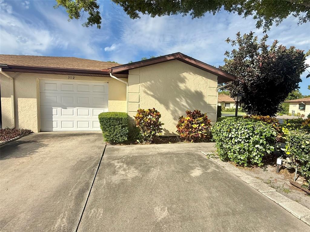 A single-story house exterior features a white garage door, beige stucco walls, and landscaped bushes — 3112 VIVIENDA BOULEVARD, BRADENTON, FL