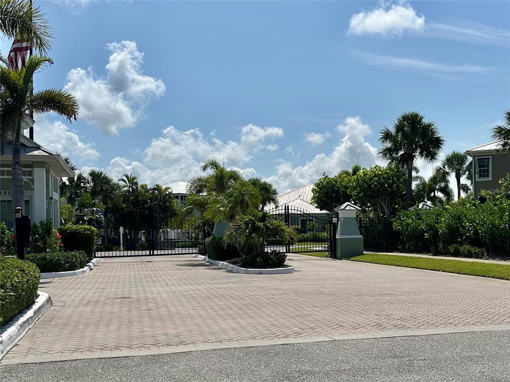 A gated entrance features a paved driveway, lush landscaping, and palm trees under a blue sky — 10600 LEMON CREEK LOOP UNIT 101, ENGLEWOOD, FL