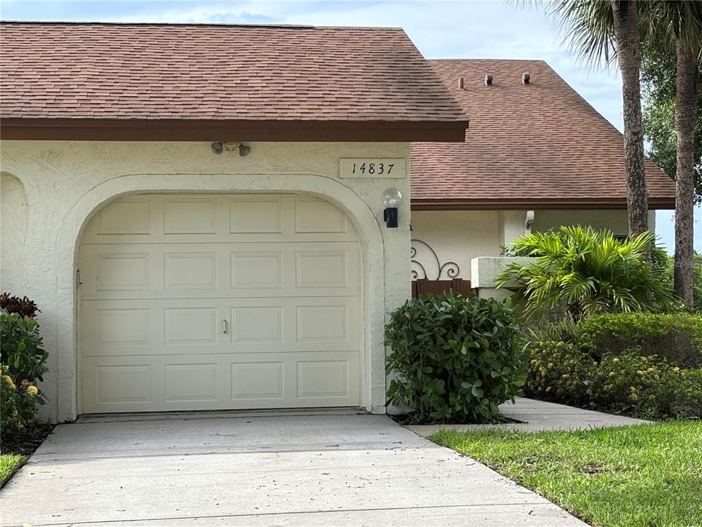 A beige garage door with an arched frame sits beside a house with brown shingles — 14837 FEATHER COVE LANE UNIT 45, CLEARWATER, FL