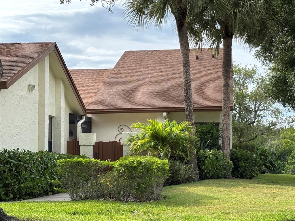 A tan stucco house with a brown shingled roof sits behind green shrubs and palm trees — 14837 FEATHER COVE LANE UNIT 45, CLEARWATER, FL