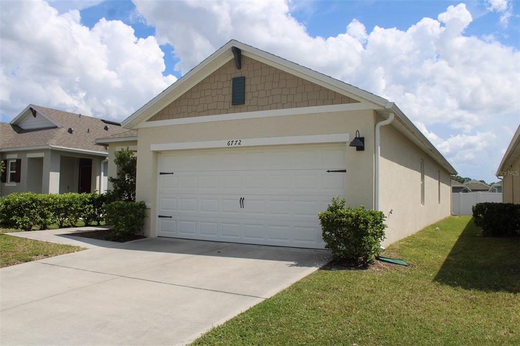 A single-story house exterior features a white garage door, tan stucco walls, and shingled siding — 6772 HUNTLEIGH HAMMOCK RD, HARMONY, FL