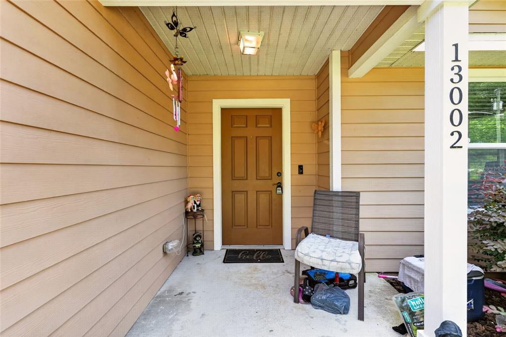 A covered front porch features tan siding, a brown door, and a single outdoor chair — 13002 SW 174TH STREET, ARCHER, FL