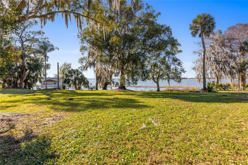 A grassy yard leads to a lakefront view with large trees and a small dock — 6136 DORA DRIVE, MOUNT DORA, FL