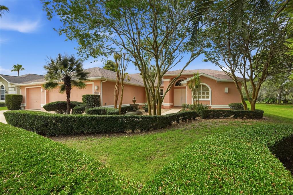 A peach-colored single-story house with a manicured hedge, a fountain, and mature trees — 4159 ST IVES BOULEVARD, SPRING HILL, FL