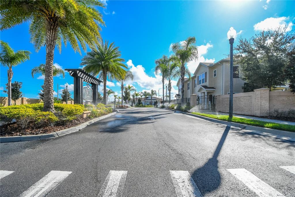 A paved road leads through a gated residential entrance lined with palm trees and landscaping — 8927 SHINE DRIVE, KISSIMMEE, FL