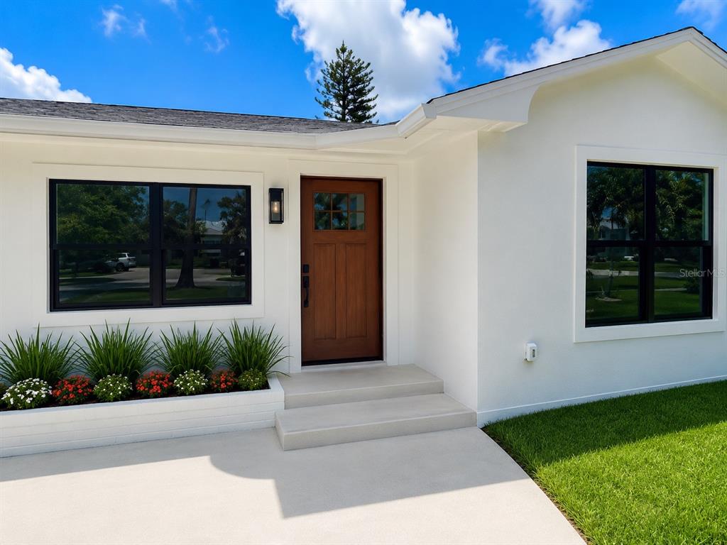 A white house exterior features a wooden front door, black-framed windows, and a landscaped planter — 3219 E DEBAZAN AVENUE, ST PETE BEACH, FL