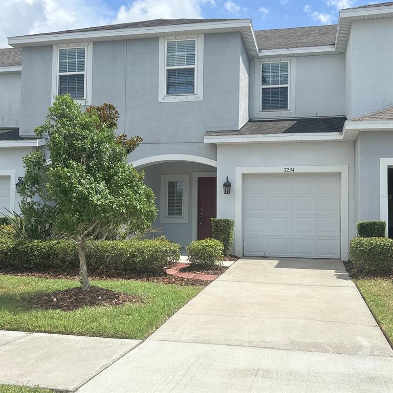 A two-story townhouse exterior features a light gray facade, a single-car garage, and landscaping — 7234 GRAND ELM DRIVE, RIVERVIEW, FL