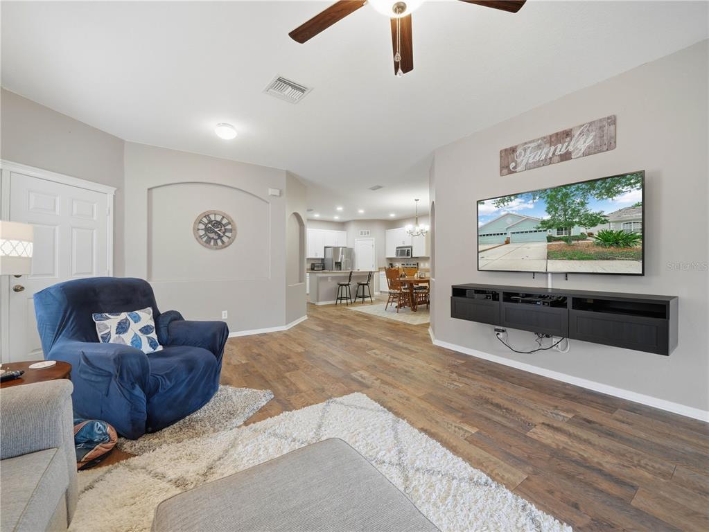 A living room features wood-look flooring, a wall-mounted television, and an open view into the kitchen — 6936 SURREY OAK DRIVE, APOLLO BEACH, FL