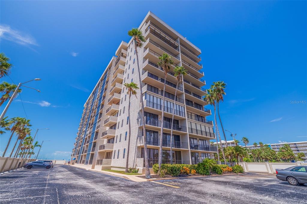 A tall, multi-story concrete apartment building stands under a clear blue sky with palm trees — 5396 GULF BOULEVARD UNIT 209, ST PETE BEACH, FL