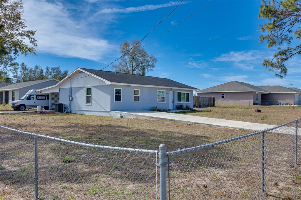 A light blue single-story house with a carport and a chain-link fence in the foreground — 9782 SE 157TH LANE, SUMMERFIELD, FL